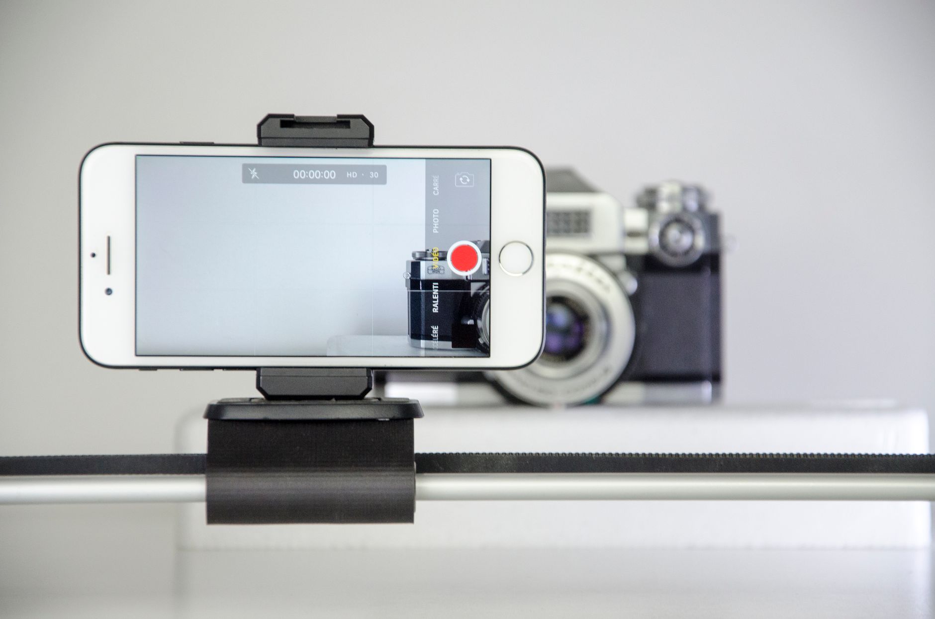 A smartphone in a clamp on a slider, its screen in video mode with the record button showing, in front of a vintage camera.
