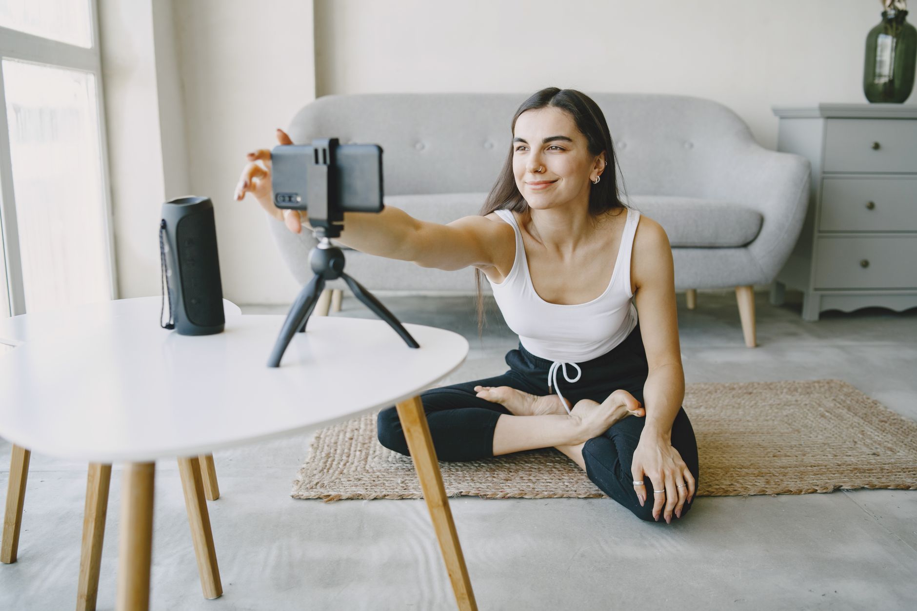 A woman sitting on a living-room floor, reaching out to adjust a smartphone on a small tripod before filming a short product video.