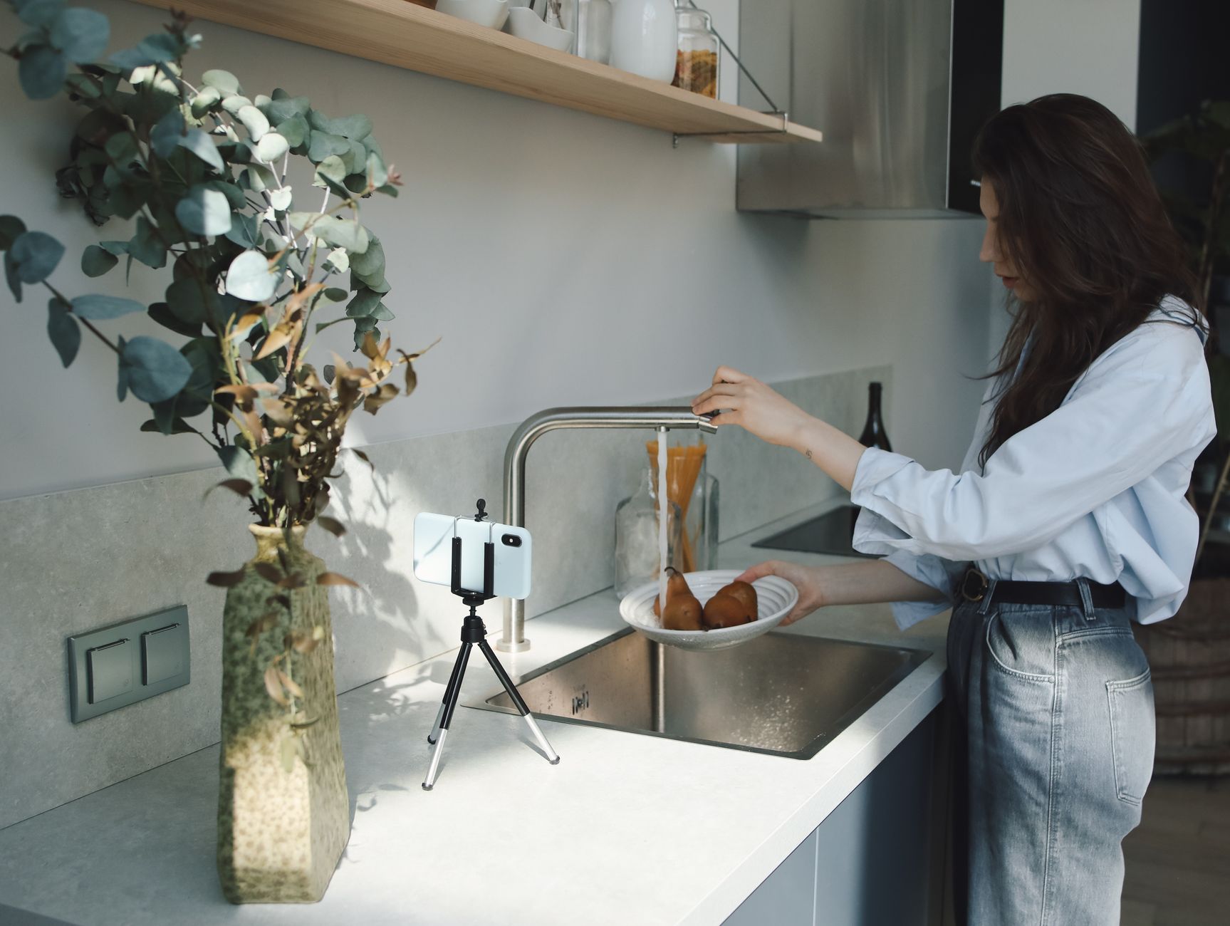 A woman rinsing pears at a kitchen sink while a smartphone on a small tripod films her from the worktop.
