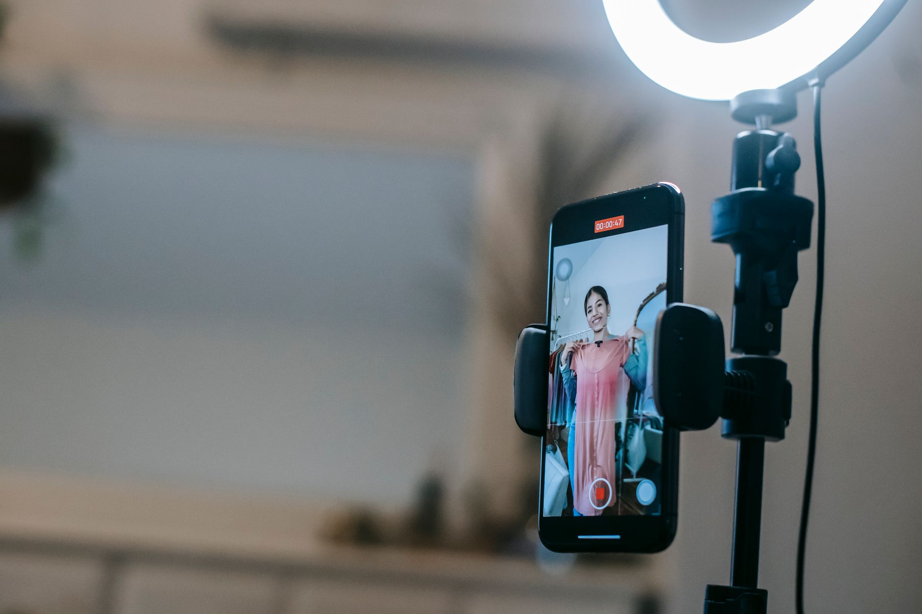 A smartphone on a stand beside a ring light, its screen showing a person holding up a pink dress while recording.